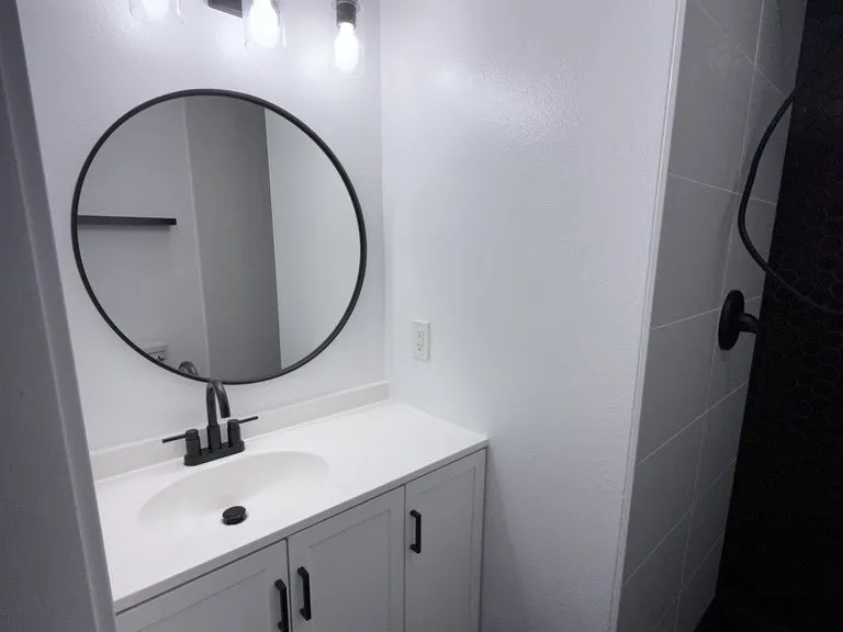 A white vanity with a round black mirror and a wall sconce