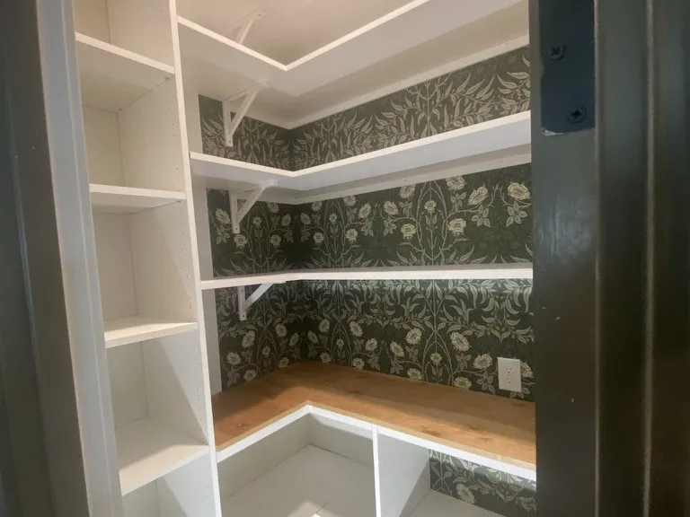 A walk-in pantry with white shelving and patterned wallpaper