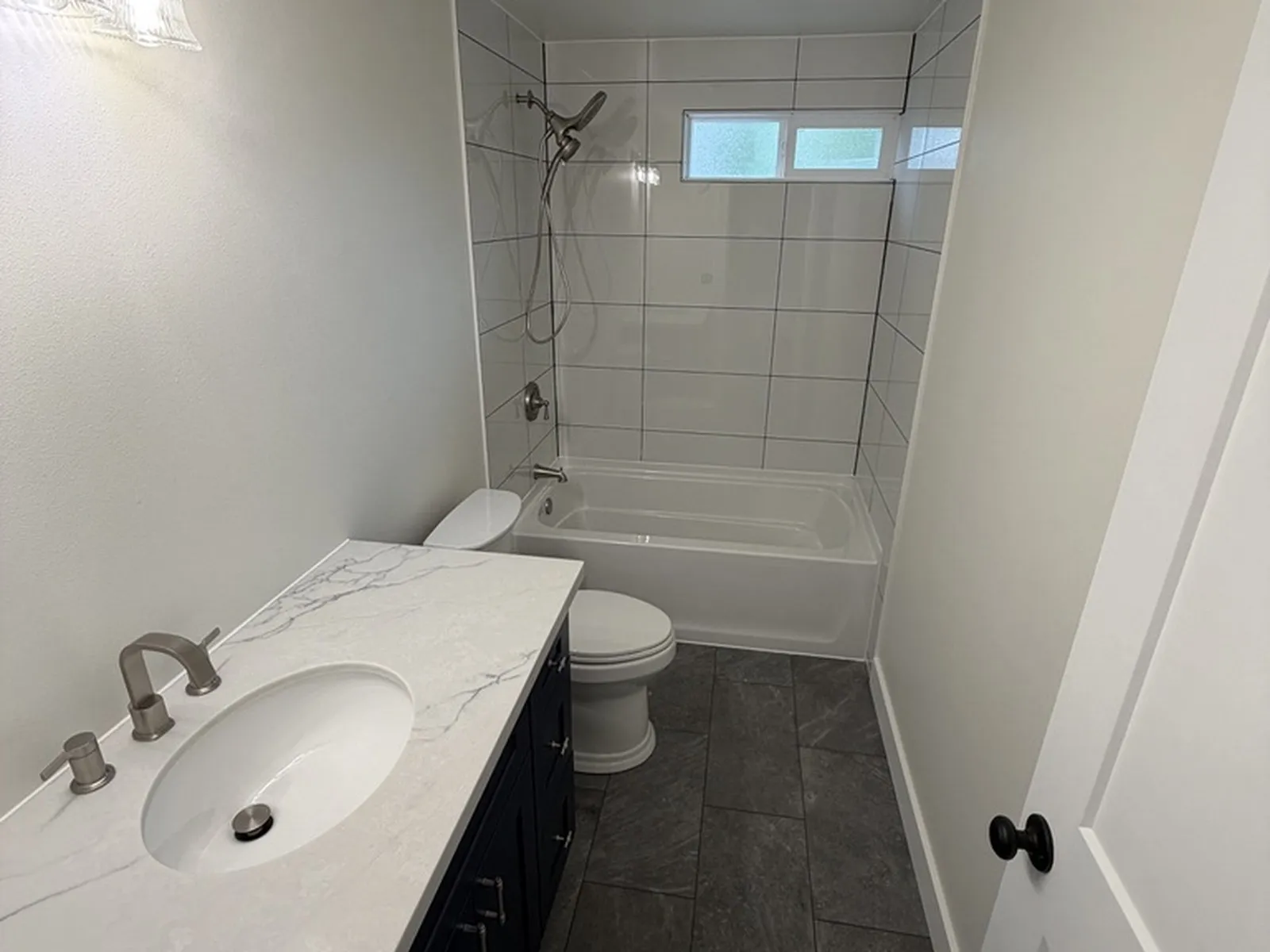 The same hall bathroom finished, with a navy vanity, a stone counter, tiled tub surround and slate-look floor tile